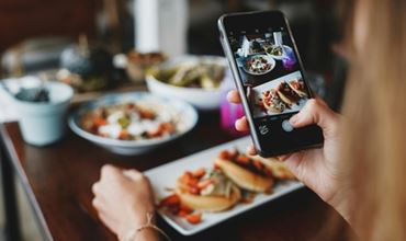 Person taking a photo of prepared dishes with a smartphone in a restaurant, with several plated meals on the table.