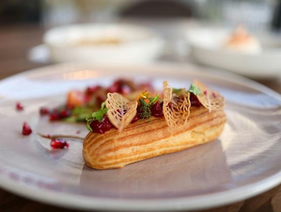 Refined éclair-style dessert topped with red fruits, decorated with crispy elements and herbs on an elegant plate.