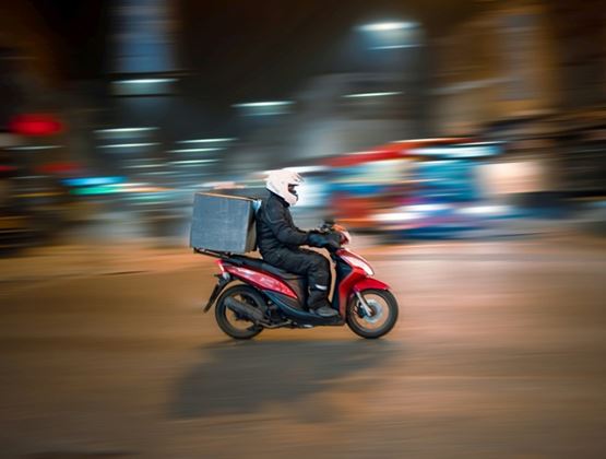 Delivery rider on a scooter speeding through a city at night, with a delivery box on the back, against a blurred urban background suggesting motion.