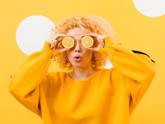 Person with curly blonde hair wearing a yellow sweatshirt, holding two lemon slices in front of their eyes against a bright yellow background, in a playful and colorful setup.