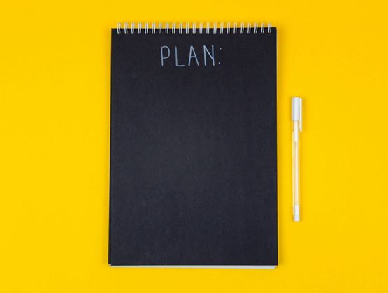 Black spiral notebook with the word “PLAN” written at the top, accompanied by a pen, placed on a bright yellow minimalist background.