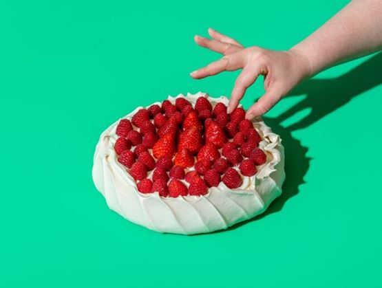 Strawberry-covered cake on a bright green background, with a hand reaching to pick a strawberry, creating a tempting and dynamic scene.