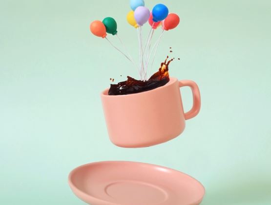 Pink cup floating above its saucer, filled with splashing coffee and decorated with colorful balloons.