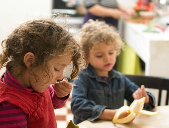 Deux enfants mangent des fruits à table pendant qu’un adulte prépare un repas en arrière-plan.