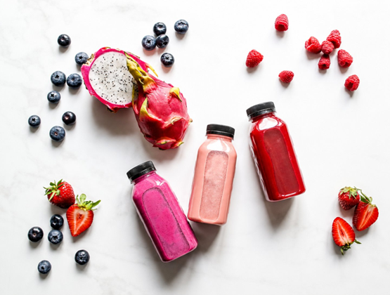 Trois bouteilles de smoothies aux fruits rouges disposées sur un fond blanc, entourées de myrtilles, framboises, fraises et fruit du dragon.