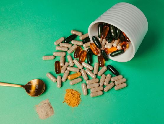 White jar tipped over, spilling dietary supplement pills and capsules onto a green background, with a spoon and powders nearby.
