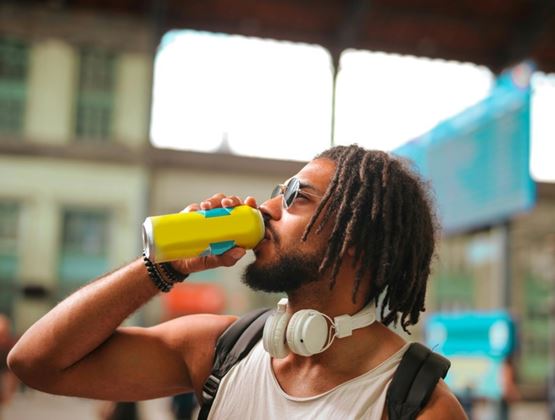 Man with headphones around his neck drinking from a yellow can in a public place.