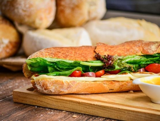Baguette sandwich filled with cheese, lettuce and tomatoes, served on a wooden board with loaves of bread in the background.