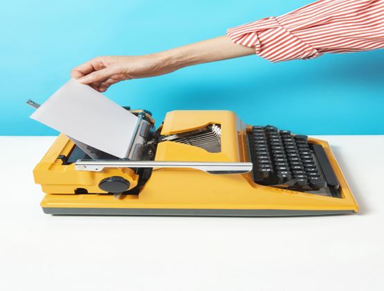 Hand placing a sheet of paper into a yellow typewriter against a bright blue background.