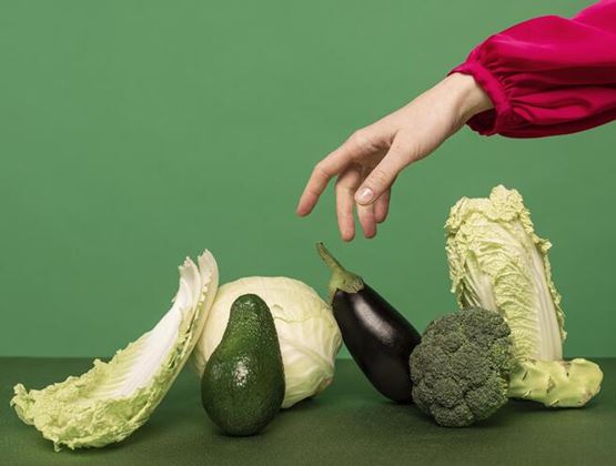 Main tendue vers des légumes frais, dont du chou, un avocat, une aubergine et du brocoli, sur un fond vert.