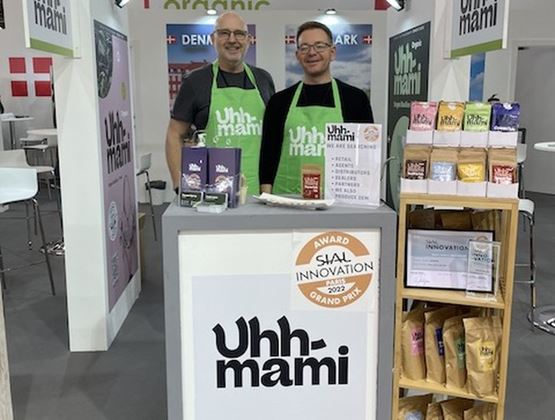 Two exhibitors wearing green Uhhmami aprons at a Danish booth at SIAL, presenting the brand’s products and promotional materials.