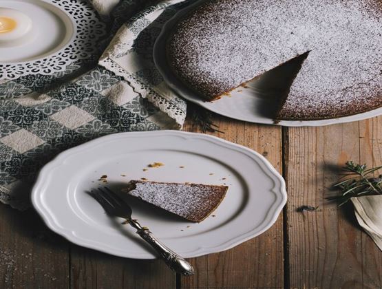 Slice of chocolate cake served on a white plate, with the remaining cake dusted with powdered sugar on a rustic wooden table.