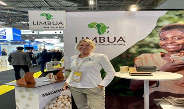 Représentante de LIMBUA devant le stand macadamia de l’entreprise lors d’un salon professionnel, avec signalétique de marque et stands en arrière-plan.