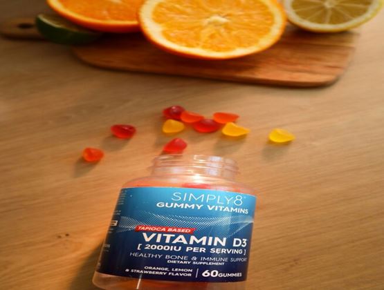 Open bottle of gummy vitamins on a wooden table with colourful gummies and sliced citrus fruits in the background.