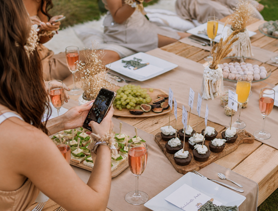 Person photographing a decorated picnic table with a smartphone, surrounded by drinks, desserts and appetisers at an outdoor gathering.