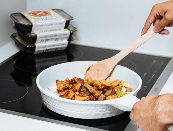 Person stirring a prepared meal in a white pan on a stovetop, with stacked food trays in the background.