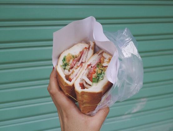 Main tenant un banh mi coupé en deux dans son emballage, devant un volet vert.