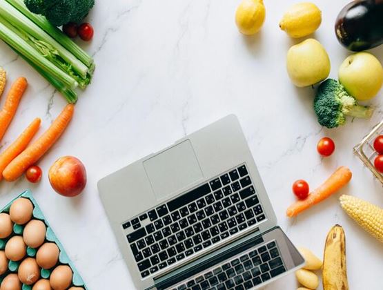 Laptop surrounded by fresh fruits, vegetables and eggs on a bright kitchen worktop.