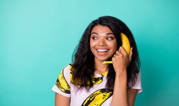 Smiling woman holding a banana to her ear like a telephone against a turquoise background.