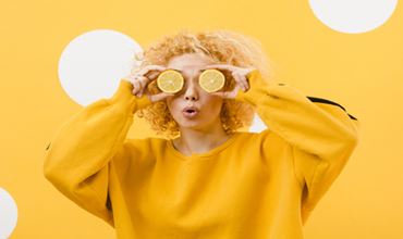 Femme vêtue d’un pull jaune tenant des rondelles de citron devant ses yeux, sur un fond jaune vif.