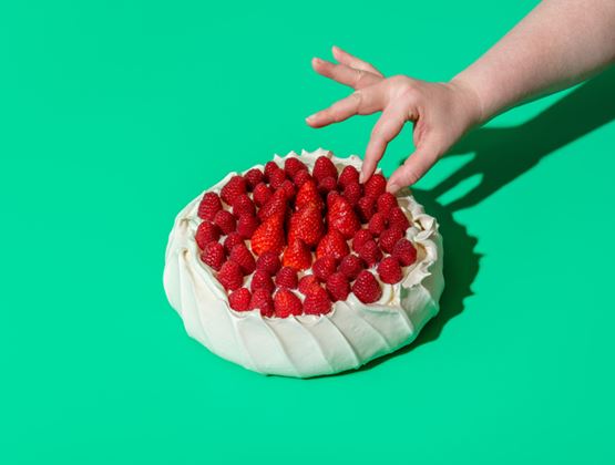 Pavlova garnie de framboises sur un fond vert vif, avec une main tendue vers le dessert.