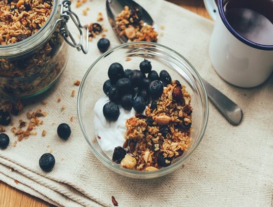 Bowl of yoghurt topped with granola, dried fruit and blueberries, beside a jar of granola and a mug.