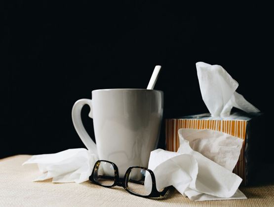 Tasse, boîte de mouchoirs, mouchoirs usagés et lunettes disposés sur une table devant un fond sombre.