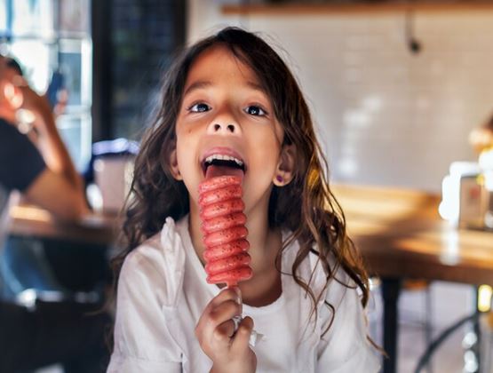 Young girl holding and tasting a red spiral-shaped ice pop indoors.