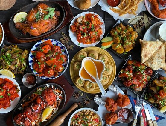 Vue de dessus d’une table garnie de plats colorés, avec des viandes grillées, des bouchées frites, des sauces, des légumes et du pain.