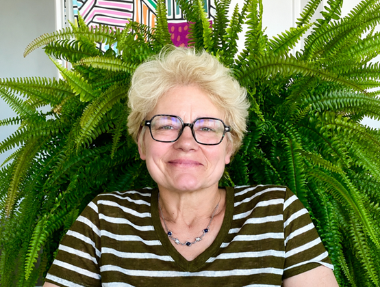 Portrait d'une femme souriante aux cheveux blonds courts et lunettes noires, devant une grande fougère verte