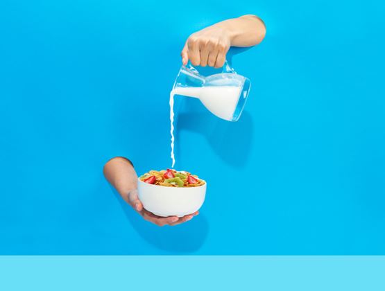 Du lait versé d'une carafe en verre dans un bol de céréales aux fraises, tenu à bout de bras sur fond bleu vif
