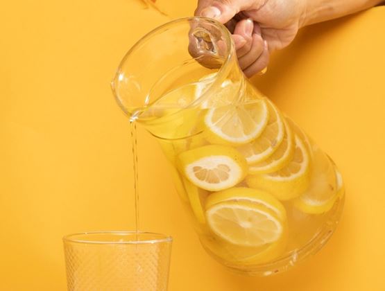 Lemon water poured from a jug filled with lemon slices into a glass, against a bright yellow background