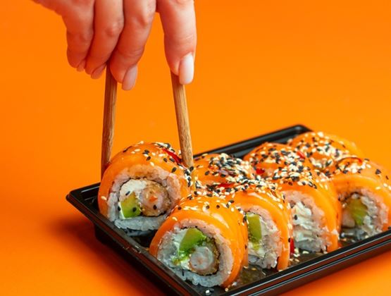 Hand using chopsticks to pick up a salmon sushi roll from a tray against a bright orange background.