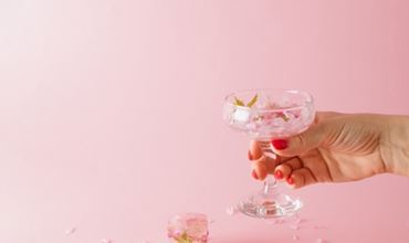 A hand with red nails holds a glass coupe filled with flower petal ice cubes, against a pink background