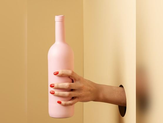 Une main aux ongles rouges tient une bouteille rose pastel, surgissant d'un mur sur fond beige