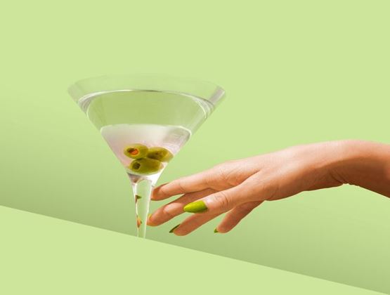 Main aux ongles verts s’approchant d’un verre à cocktail contenant trois olives, sur fond vert clair.