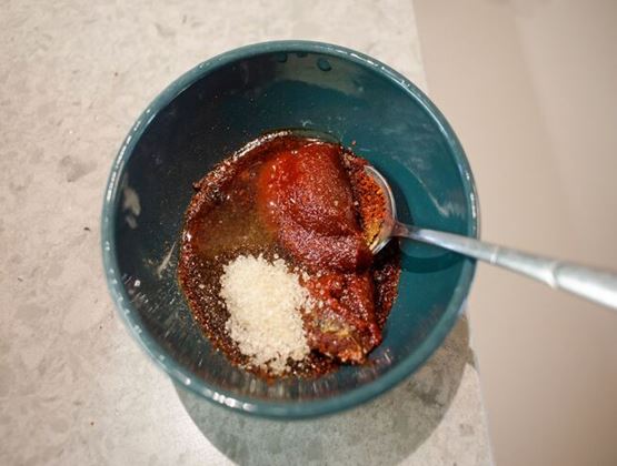 Bowl containing tomato paste, sugar and spices being mixed with a spoon.