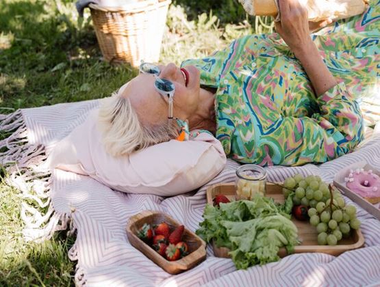 Femme âgée allongée sur une couverture lors d’un pique-nique, entourée de fruits, de légumes et d’un panier en osier.