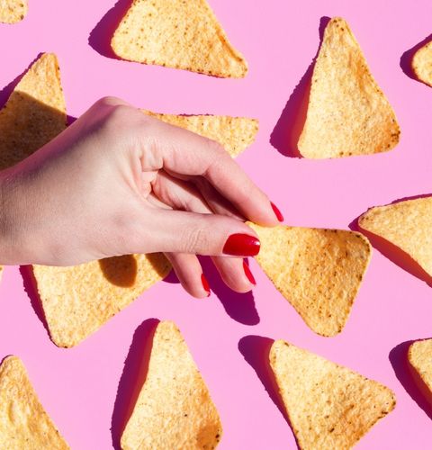 Hand with red nails holding a triangular chip among others, on a bright pink background.