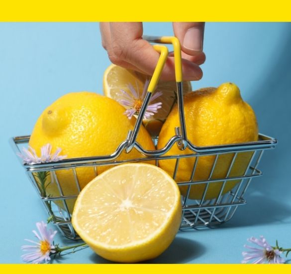 Hand holding a small metal basket filled with yellow lemons, with a half lemon in the foreground on a blue background.