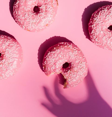Donuts glacés roses avec vermicelles blancs, dont un croqué, disposés sur fond rose avec ombre de main.