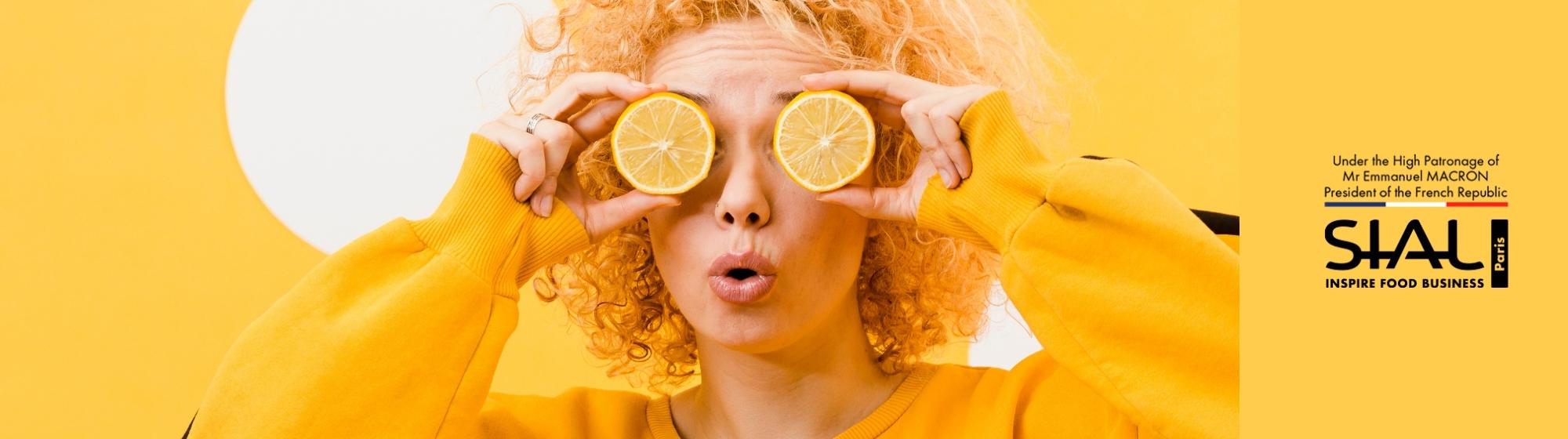 Curly-haired woman holding two lemon slices over her eyes on yellow background with SIAL Paris logo and official mention
