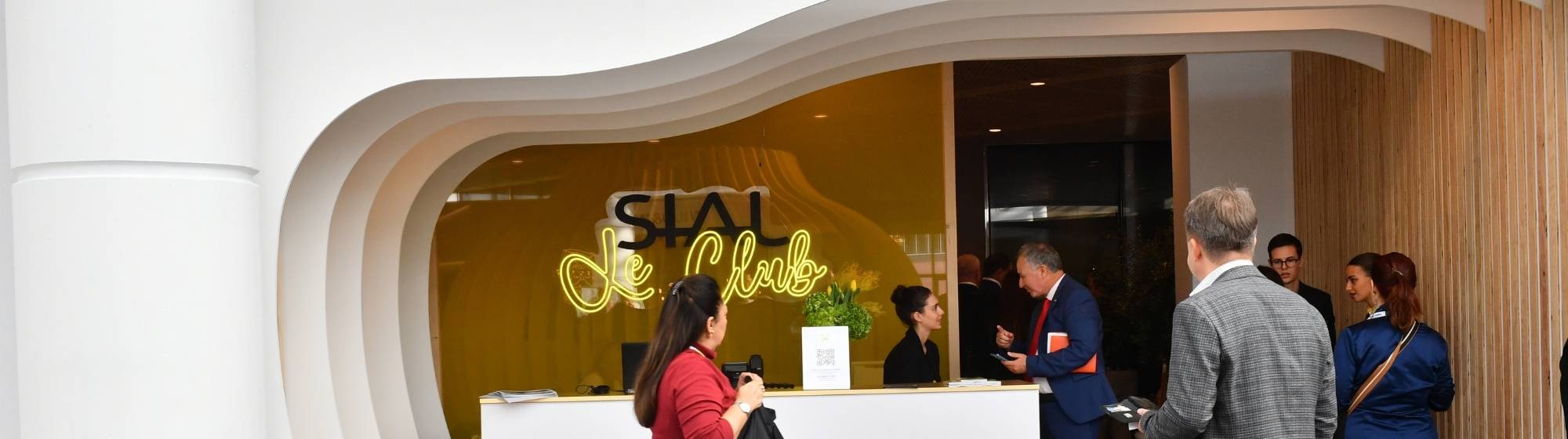 Accueil du SIAL Le Club avec visiteurs et personnel discutant devant un comptoir moderne et lumineux.