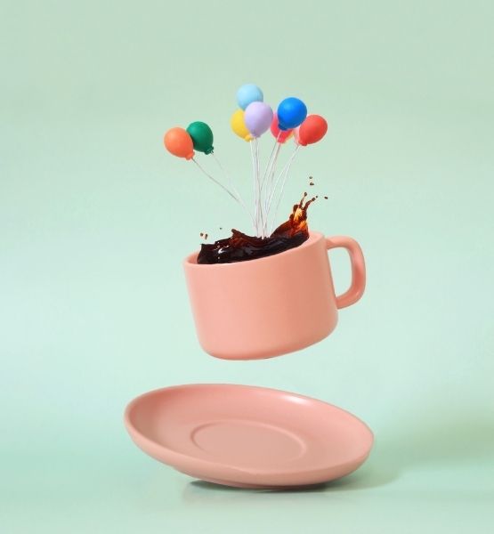 Tasse rose flottante avec éclaboussure de café et petits ballons colorés sur fond vert pastel.