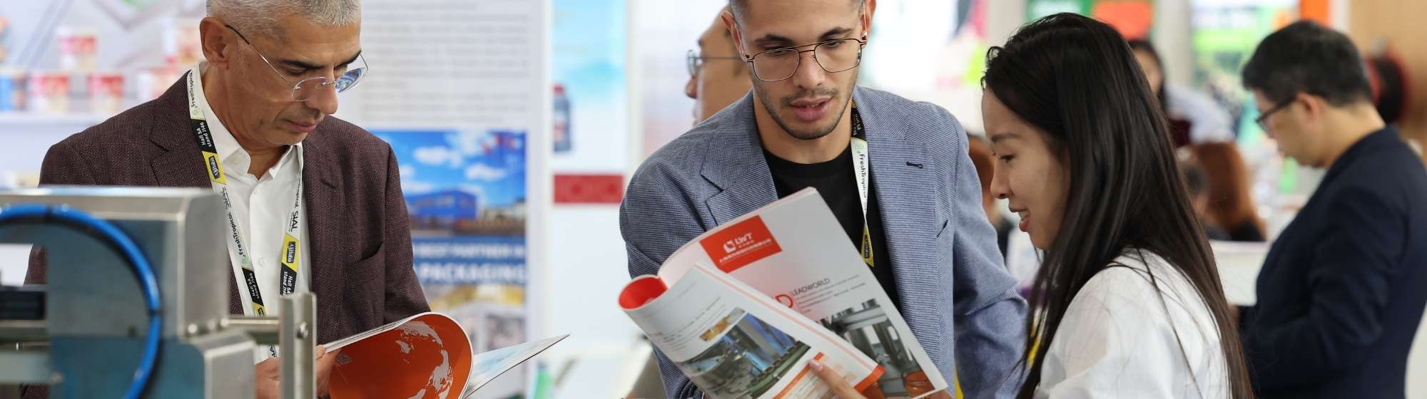Trois visiteurs consultent une brochure sur un stand présentant des équipements de conditionnement.