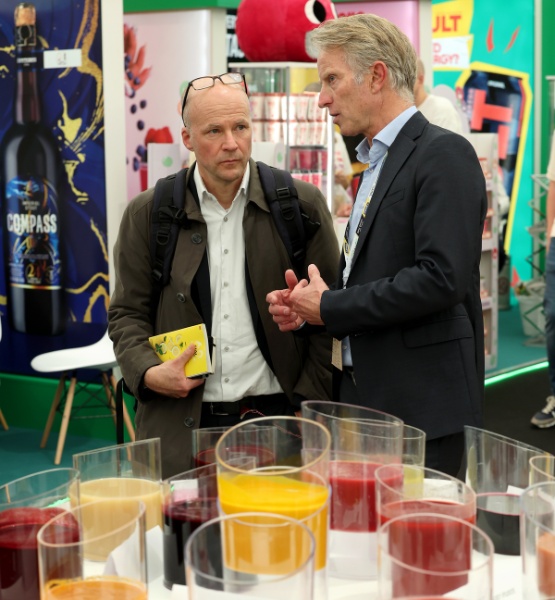 Two visitors talk in front of a display of colored glasses at a trade show booth.