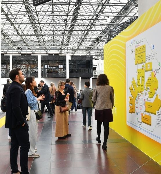 Visitors checking a trade show map on a large yellow wall in an exhibition hall.