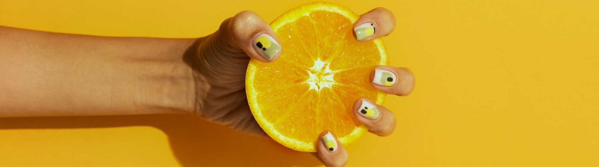Hand firmly holding half an orange against a bright yellow background, with original patterns on the nails.