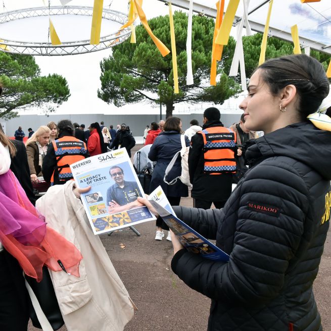 Personne distribuant des brochures à l'extérieur de l'événement SIAL Paris, avec des participants en arrière-plan