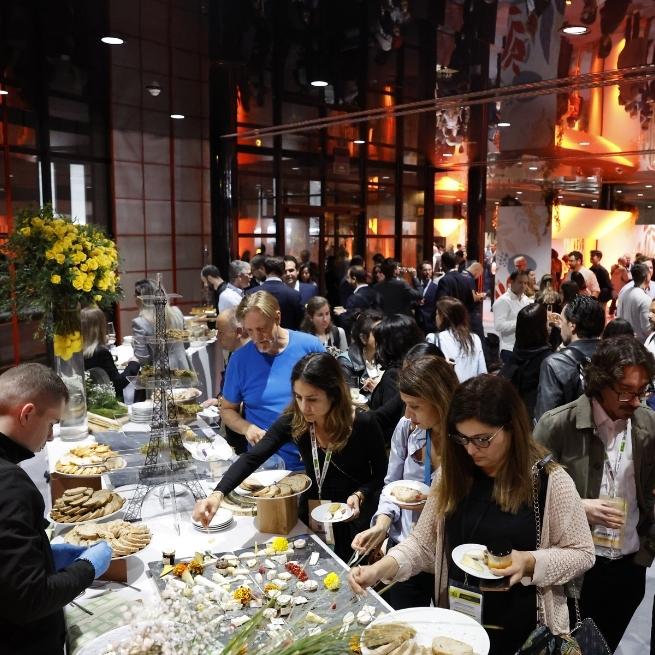 People serving food at an event with elegant decoration and flowers, surrounded by a crowd in a hall.